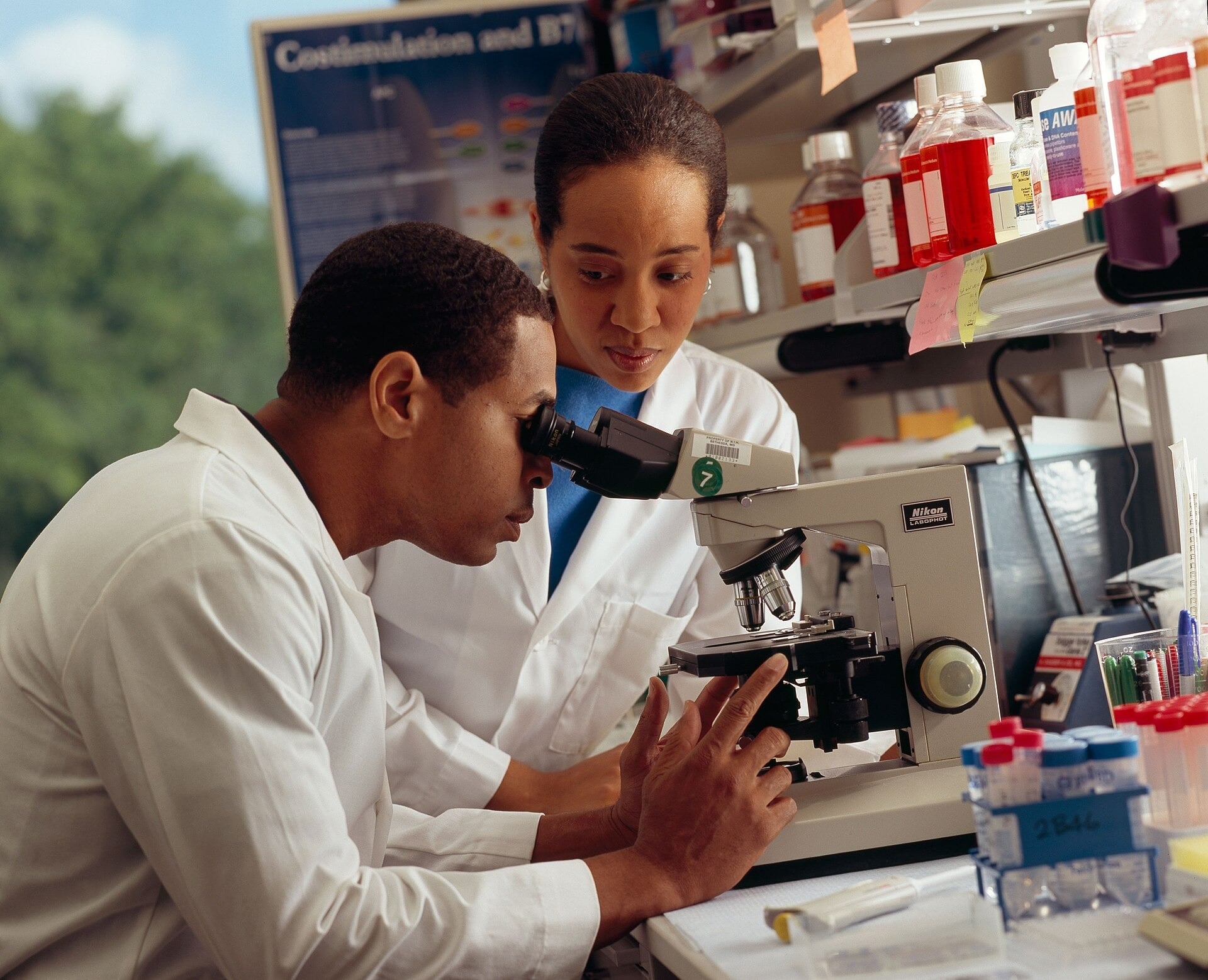 Two researchers in white lab coats work at a laboratory microscope, one looking through the eyepieces