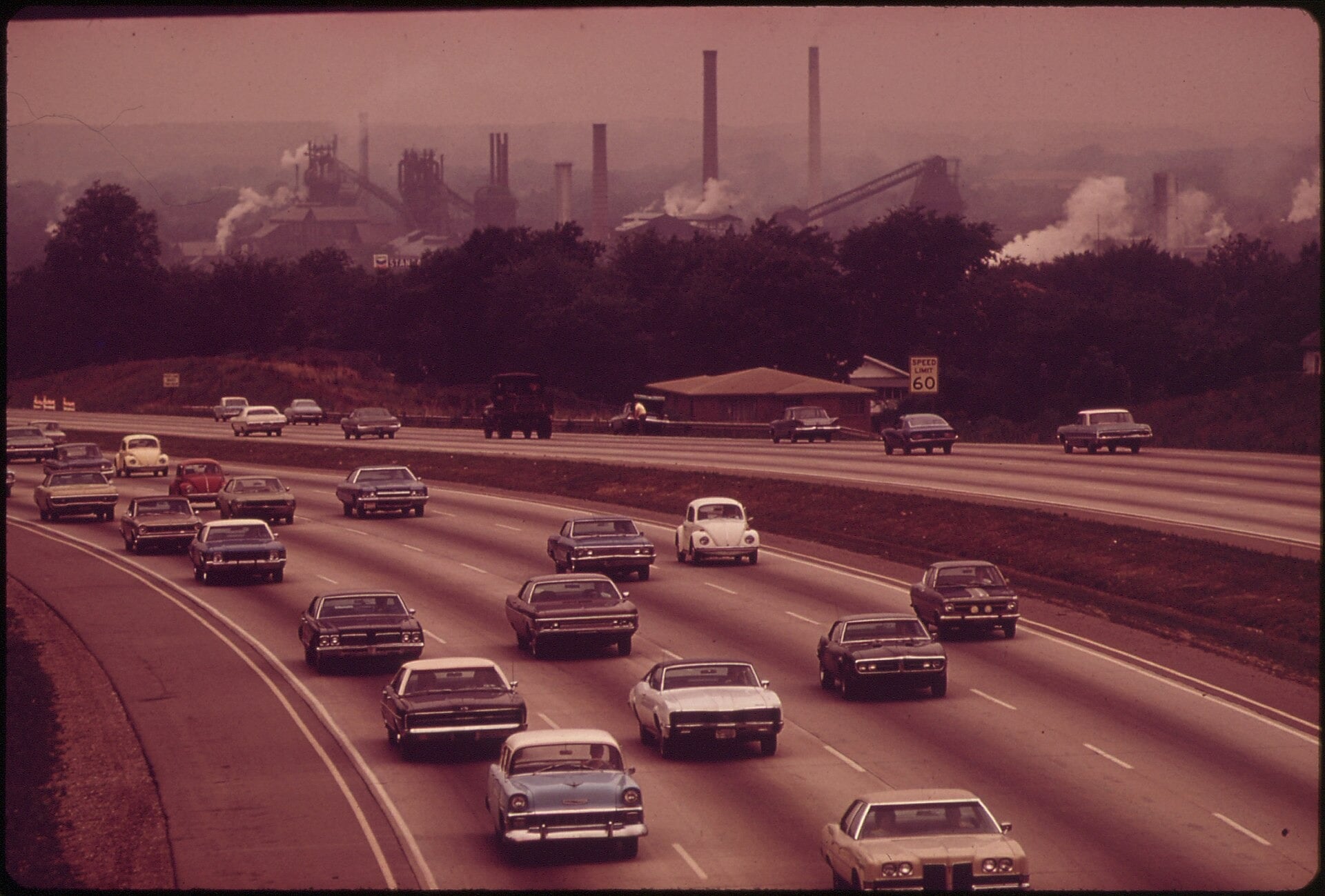 A 1970s highway full of cars in front of an industrial skyline with smokestacks releasing smoke into hazy air