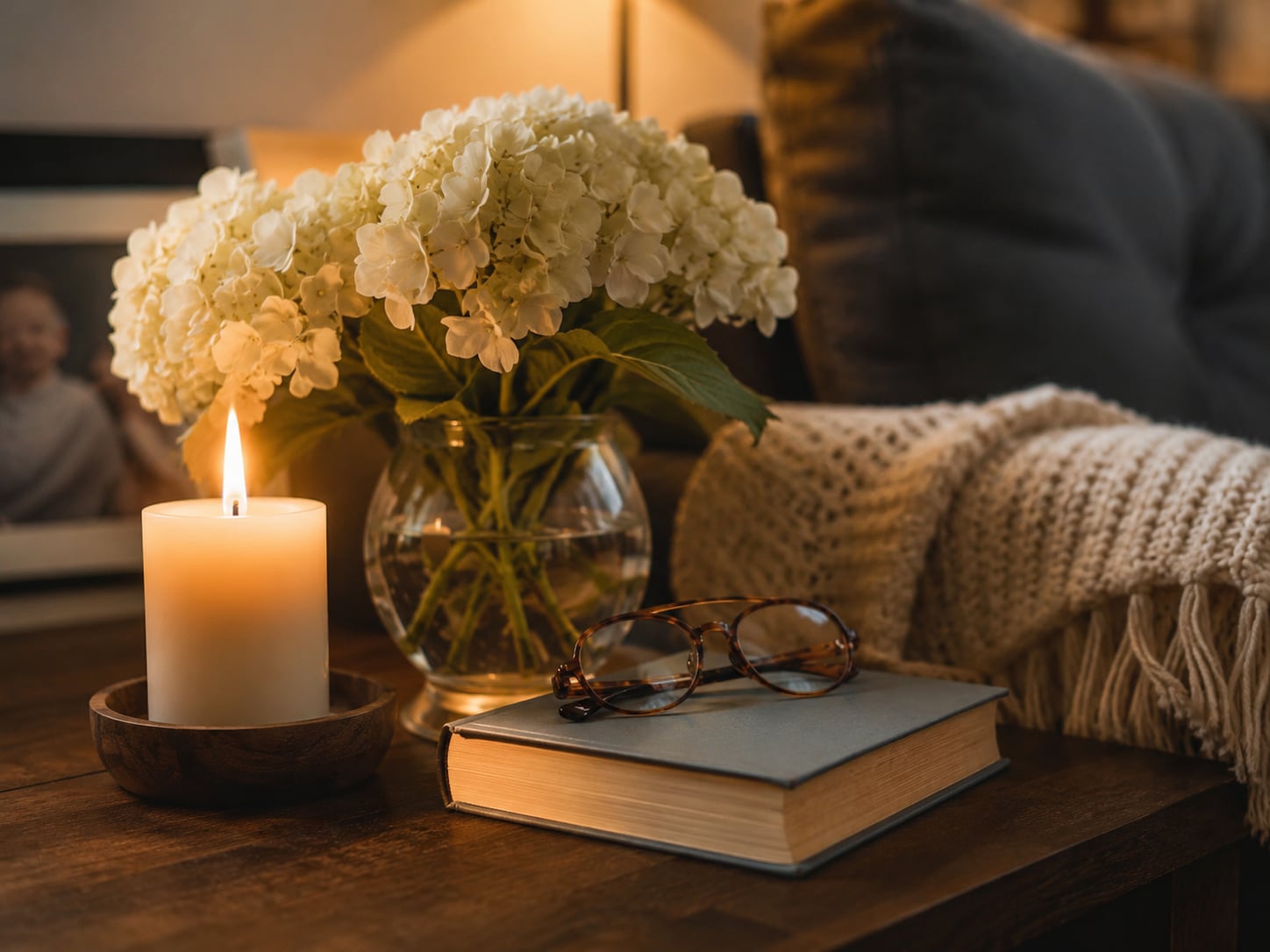 A lit pillar candle, a vase of white hydrangeas, and reading glasses resting on a closed book on a wooden table beside a knitted throw.
