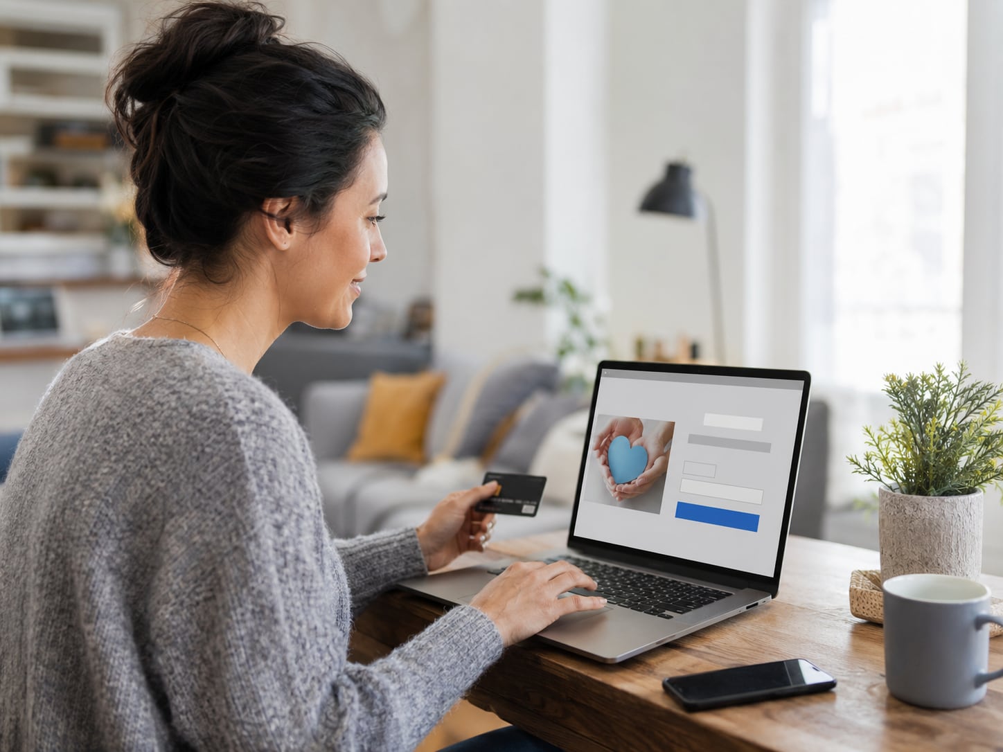 A woman sits on a couch using a laptop and a credit card, appears to be paying online