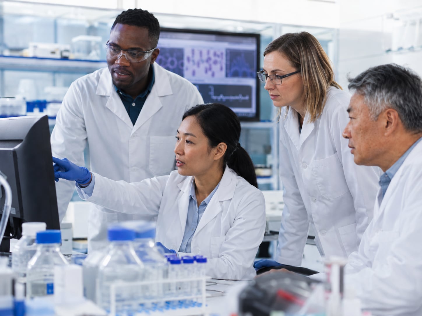 Three lab researchers in coats examine samples together at computer monitors in a laboratory