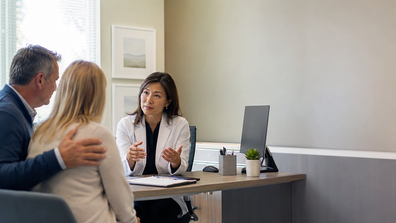 A female doctor talks with an older man, hand on his shoulder, in an office setting
