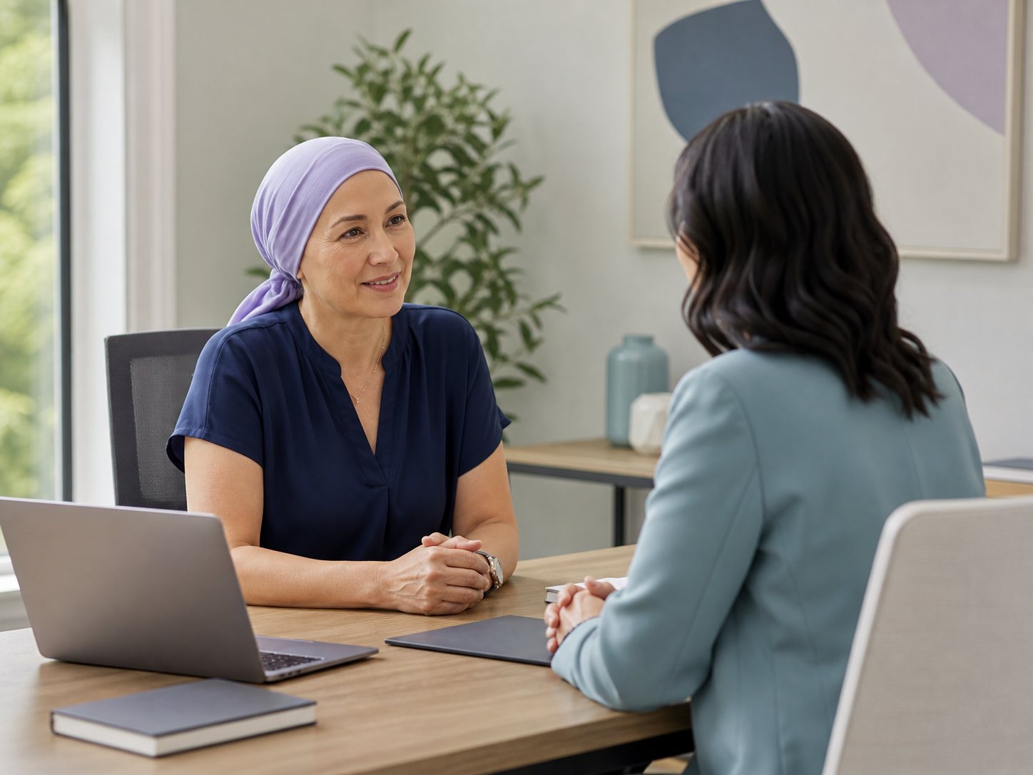 A woman in a headscarf sits across a desk from a colleague with an open laptop, in an unbranded workplace.