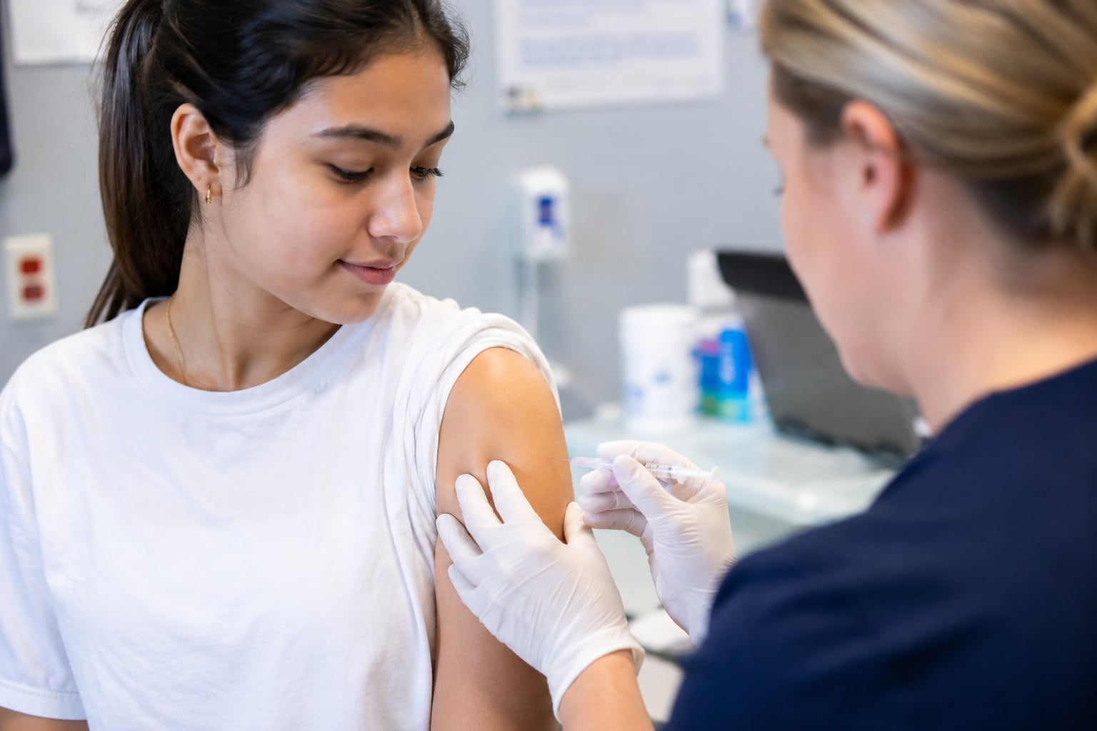 A clinician in navy scrubs and white gloves holds a syringe at a young woman’s bared upper arm in a clinic room.