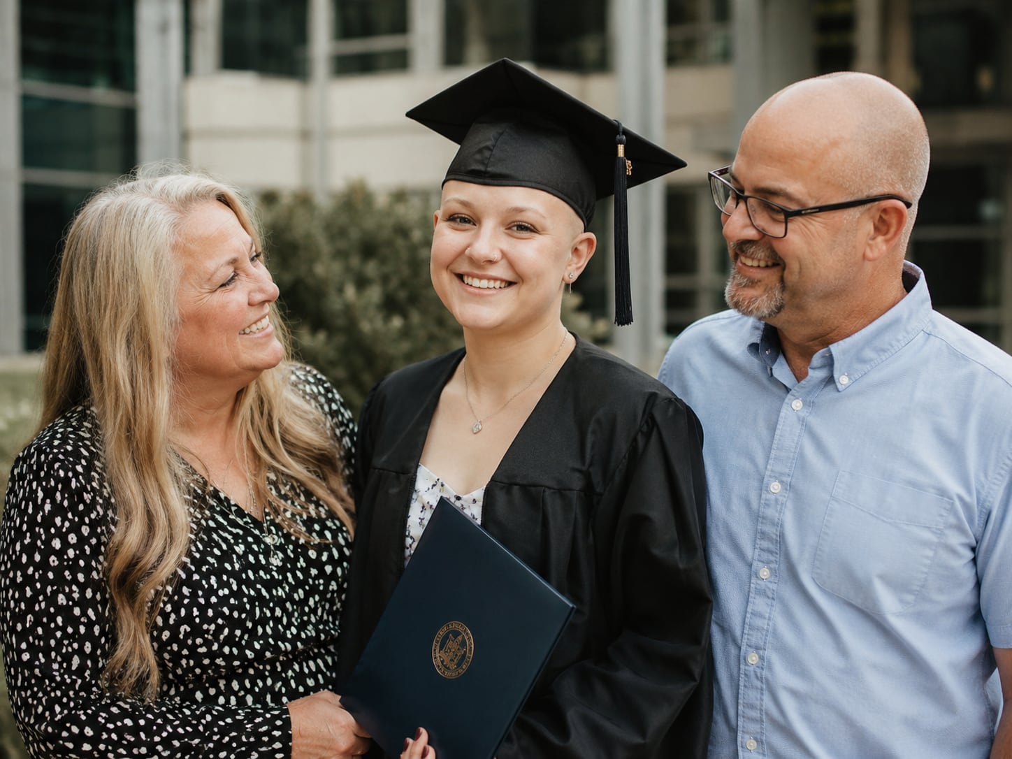 A young woman with a bald head in a graduation cap stands with two smiling parents